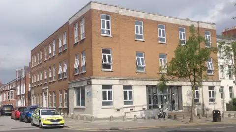 The corner view of a building - it is red brick on top and white brick on the bottom. A police car is at the side of the building.