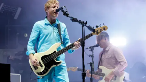 Jamie Simonds/BBC Two band members playing guitars and singing into microphones on stage at Reading Festival. One is wearing a bright blue tracksuit jacket and trousers, the other is wearing a pale pink shirt.