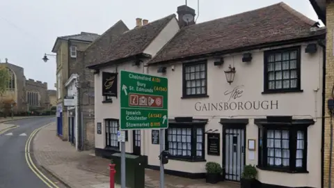 A general view of the Gainsborough pub in Sudbury. It is beside a road and is a white building with the name of it painted on the front. A large green road sign is placed in front of it.