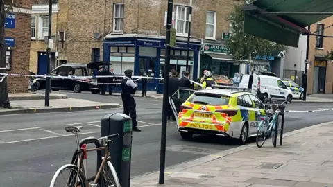 Police car surrounded by police tape, officers and the open boot of a black funeral car