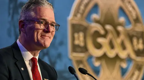 Getty Images A man is at a podium with microphones. He is wearing a black suit, white shirt, red patterned tie and has glasses and short grey hair. He has a small smile on his face. In the background is an image of the GAA logo.