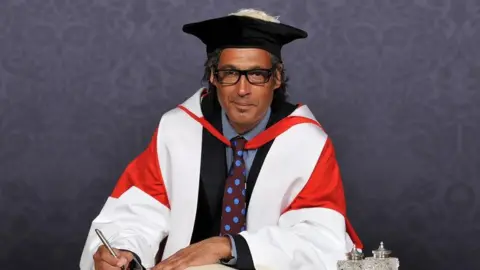 David Olusoga smiling into the camera. He is wearing black glasses, a black graduation cap and red and white robe. Underneath the robe he is wearing a dark suit with a maroon and blue spotty tie. He is writing in a book and sitting in front of a blue, patterned background.