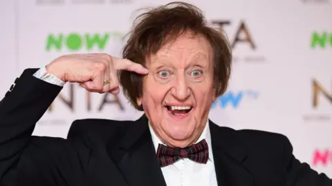 PA Media Dodd at an awards ceremony in 2017. He smiles, opened-mouth with his finger points to his temple.