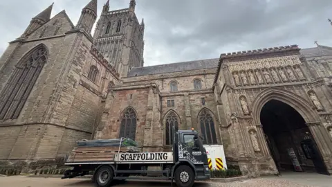 Worcester Cathedral dominates the frame, with intricate Gothic stonework and spires beneath a grey sky. A scaffolding truck is parked outside the entrance.