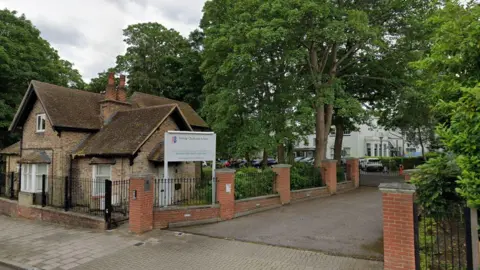Google maps picture of Bishop Challoner School. It has a series of brick posts going along a driveway and trees to the side. The school building is small with a pitched roof and there is a white school sign out front.