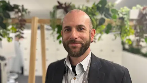 Phil Smith looking into camera. He has a beard and is wearing a navy suit, white shirt and a lanyard