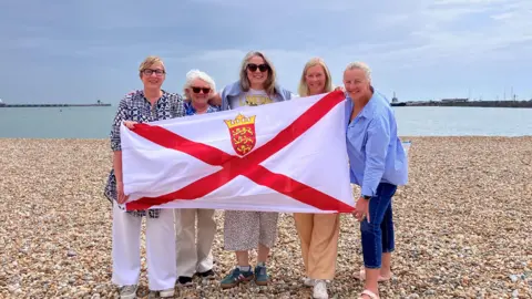 A picture of the five women standing on a beach with a flat sea behind them. They are holding the Jersey flag.