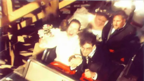 Korey Kieptfer Korey and his wife in the front cart of a rollercoaster. He is wearing a black suit, she is wearing a white wedding dress and holding a bouquet. In the row behind is another man in a dark suit and a woman in a light dress with a flower headband. 