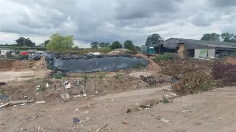 Mounds of waste, including tyres, lie on a bare patch of ground next to a large barn. 