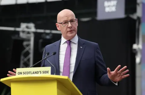 EPA John Swinney, who is bald with glasses, holds a manifesto booklet in his right hand. He is wearing a business suit with a purple tie 