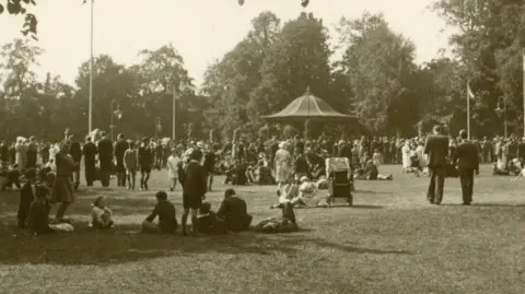 A black and white photo of people in a park. There is a bandstand in the distance with a conical roof. Some people are sitting on the grass while others are walking around. There is a large group of people near the bandstand. There are trees in the background.