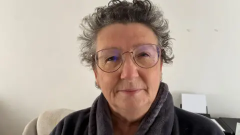 Robby West/BBC Linda Pendergrass is wearing glasses and a dark grey fleece. She is looking directly at the camera and is in front of a white wall.