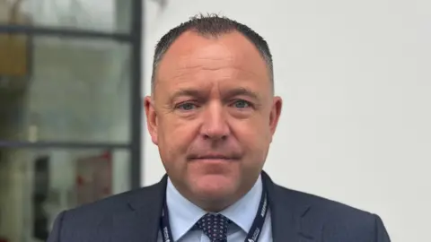 George King/BBC A head and shoulders image of Matt Adams. He is wearing a navy suit jacket over a light blue shirt with a tie. He is looking directly into the camera. 