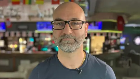Anthony Cassidy is stood in his bar looking straight at the camera. He is wearing a navy T-shirt and black glasses.