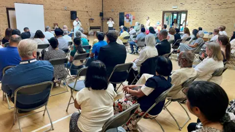 Stephen Huntley/BBC People sit in rows of chairs within a community hall. They face towards a woman speaking behind a microphone. The picture is taken from a back corner of the hall.