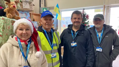 A woman wearing a white jumper and a Santa hat, stood next to three men, all of them are wearing tags which say 'Hereford Help for Ukraine'. Behind them are shoeboxes wrapped in Christmas paper. 