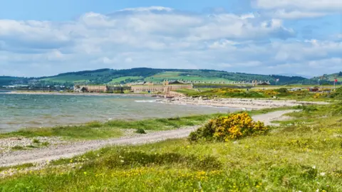 Getty Images The shoreline of the Inner Moray Firth near Ardersier. A narrow stony path and grass lines the shore. Fort George can be seen in the background.