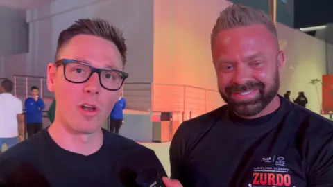 Two men in black T-shirts looking at the camera and talking.