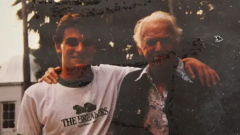 Nick Weston in a t-shirt and Douglas Fairbanks Jr in a floral shirt and jacket posing for a photograph in a garden. They have their arms around each other and are smiling.