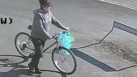 A CCTV still of a woman with dark hair, wearing a grey hooded top and dark trousers, pushing a bicycle with a blue bag on the handlebars