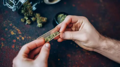Man's hands twisting marijuana into brown paper. The hands are resting on a dark table and there is a mason jar on its side with marijuana falling out of it.