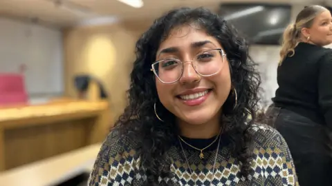 A woman with long dark hair and waiting glasses, a gold necklace and a jumper with a diamond shaped patterns is sitting in front of a row of desks.