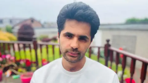 Barun Lamichhane, a man in a white T-shirt, with black hair, looking solemnly at the camera. There is a garden with flowers behind him.