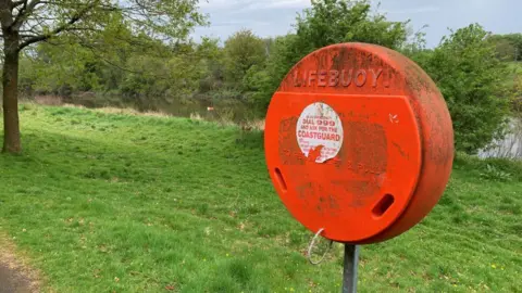 A circular, orange container marked LIFEBUOY. It is dirty and has a sticker on it advising people to call 999 for the coastguard. It is next to a grassy verge and there is a river visible beyond. 