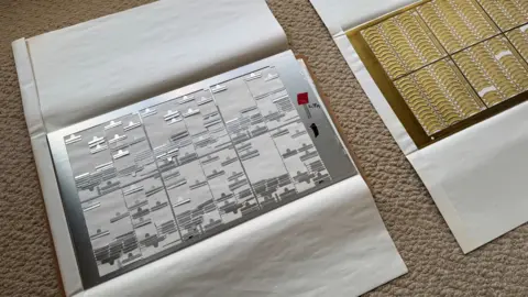 On the floor are two sheets of metal, one brass and in the foreground a stainless steel sheet. The sheets contain hundreds of raised pieces of locomotive nameplates