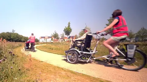 Two adapted trikes are being cycled away from the viewer along a path which is winding through a field of flowers and young trees. In the distance are some houses. The trikes are much longer than a normal bicycle and large enough on the front to accomodate a wheelchair.