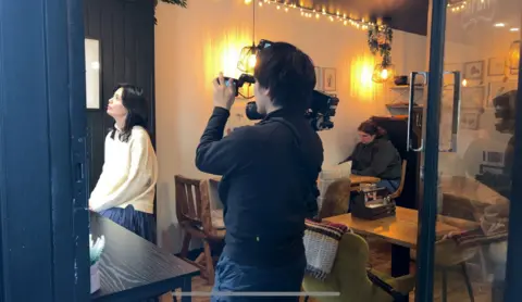 Woman in cream Guernsey getting filmed by a camera crew in the Bean Jar a local Guernsey cafe