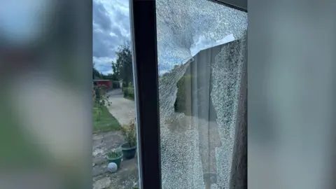 A double glazed window that is smashed and shattered. The picture is taken from inside a home. It is in a white UPCV frame and outside is a well kept garden on an overcast day.
