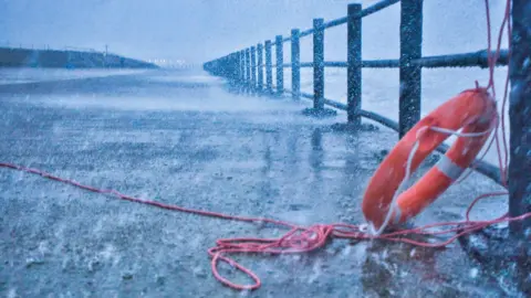 BBC Weather Watchers / Sand Dancer The promenade at Sunderland is slick with water and splashes as droplets come from the ocean beyond the grey, metal railings on the right. There is an orange life ring tied to the railings.