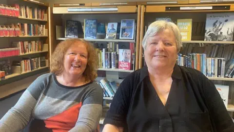 Fleetwood library users Anne and Janet sitting next to each other smiling. There are bookshelves full of books in the background. Anne is wearing a grey top with red and dark grey shapes on it and Janet is wearing a black shirt. Anne has mid-length red hair and Janet has short grey hair.