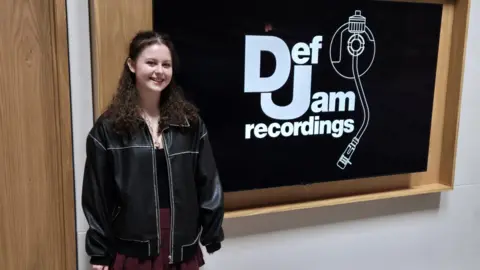 Kris Ross-Osborne Erin Rose stood in front of a sign for Def Jam Recordings. She has long dark curly hair and is wearing a black leather jacket, a black top and a burgundy skirt. She is looking at the camera and smiling. 
