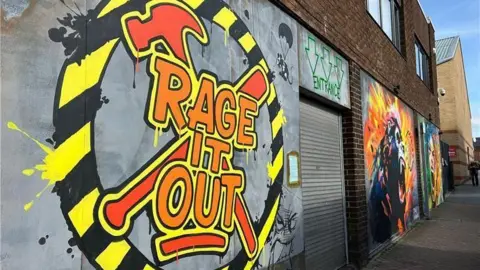 The outside of the venue, which has a graffiti sign on the wall outside with Rage It Out written in orange letters on top of a hammer and baseball bat drawing. A black-and-yellow circle surrounds the writing. To the right is a corrugated metal shutter with the word 'entrance' written in green above it. On the right of the door is another, large bit of street art.