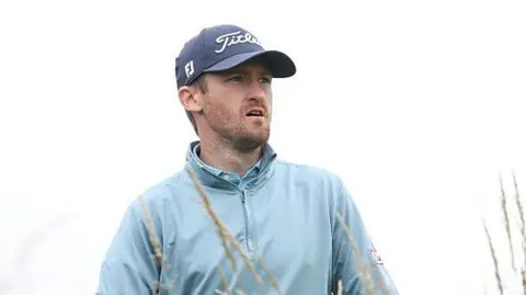 Andrew Wilson playing in The Open Final Qualifier at Dundonald Links, watching his ball as he wears a sky-blue wind-jammer 
