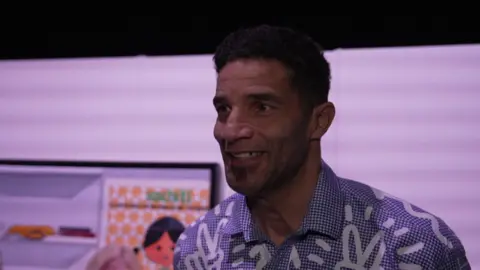 Former goalkeeper and current artist David James, smiling and looking off-camera. He has short dark hair and a blue patterned shirt. 