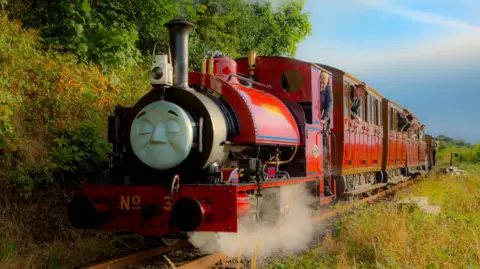 Nathan Spence A small red steam locomotive steams along the line through countryside. It has a face attached to the front, making it look like one of the characters from Thomas the Tank Engine. People in 1950s costume look out of the windows of the carriages behind.