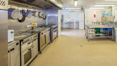 Large, clean commercial kitchen with stainless steel appliances, including multiple ovens and stovetops beneath a wide ventilation hood on the left. Pots and pans hang above the cooking area, while prep tables and sinks line the right side.