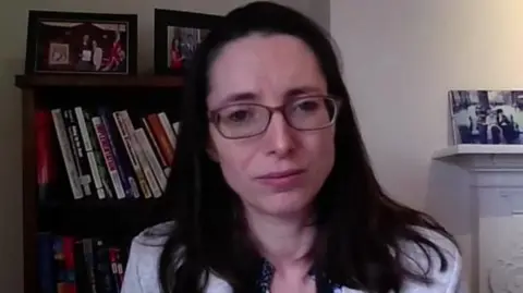 A woman with shoulder-length dark hair and glasses, wearing a grey suit jacket talking into a camera from her home. In the background is a book shelf and fireplace.