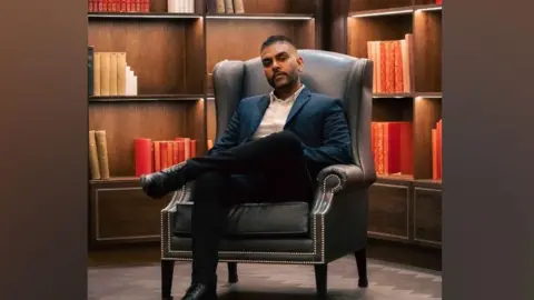San B A man with a beard sitting on a chair teal cushioned armchair. Behind him is a bookshelf with books that follow the same colour scheme of red, cream and orange.
He is not smiling at the camera, but looking directly at it 