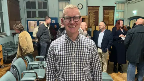 Man in checkered shirt stood infront of crowd of people in a town hall