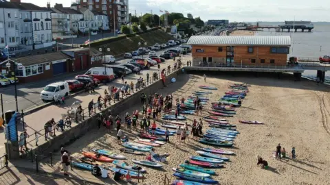 Aps Drones A group of about 30 to 40 people gathering on the seafront next to the RNLI building, with the different coloured paddleboards laid out on the beach in two rows.