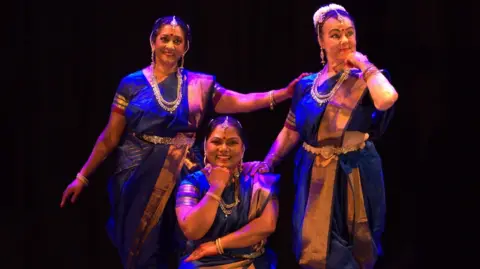 Graham Day Three performers wearing royal blue and gold Bharatanatyam costume sets. two are standing and one crouches on one knee. All are under stage lighting with a black background.