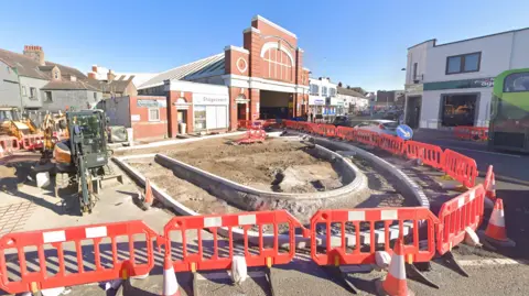 Google The street is undergoing works. Red barriers are erected around ground that is half exposed and half concreted over. There are work vehicles parked up behing the barriers. In the background is the red-brick bus building with a sign on it reading: "Stagecoach".