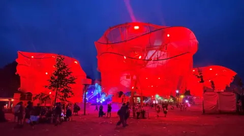 Three large paper trees lit up in red in the Shangri-La area of Glastonbury Festival.
