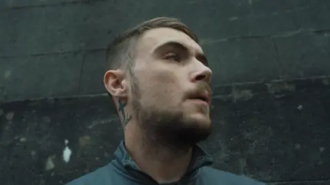 Rory Carson Pete looks to his left. He is leaning against a gray concrete wall, dressed in a dark green fleece. He has short brown hair, a short brown beard and a cross tattoo on his neck behind his right ear. 