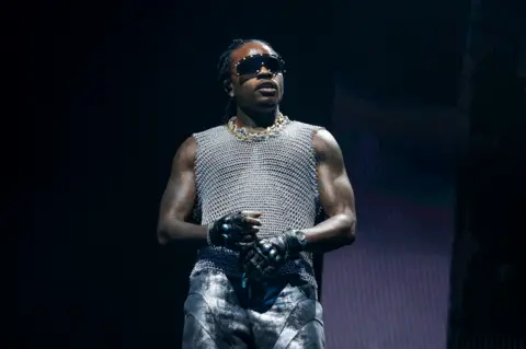 Getty Images Gunna, dressed in gold and silver chains wears a silver vest top, black gloves and sunglasses while holding a mic