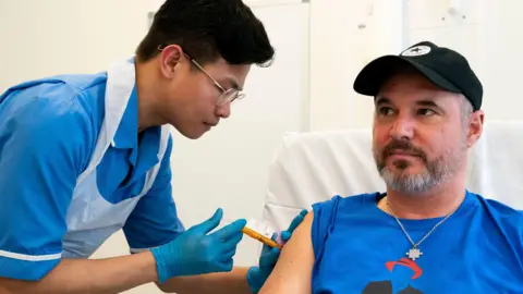 PA Media A male nurse wearing a blue uniform and white plastic apron is putting an injection into the arm of Steve Young, who is wearing a blue T-shirt and black baseball cap.